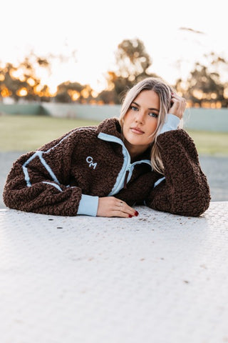 Woman wearing the Emmie Fleece in dark brown with light blue trim, leaning on a table outdoors