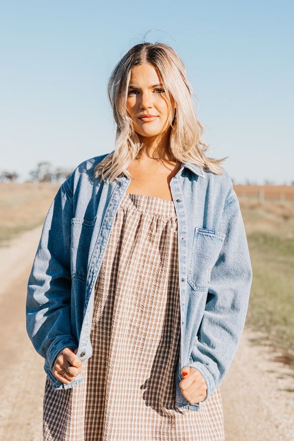 Woman wearing the Lottie Denim Shirt open over a brown gingham dress on a rural dirt road