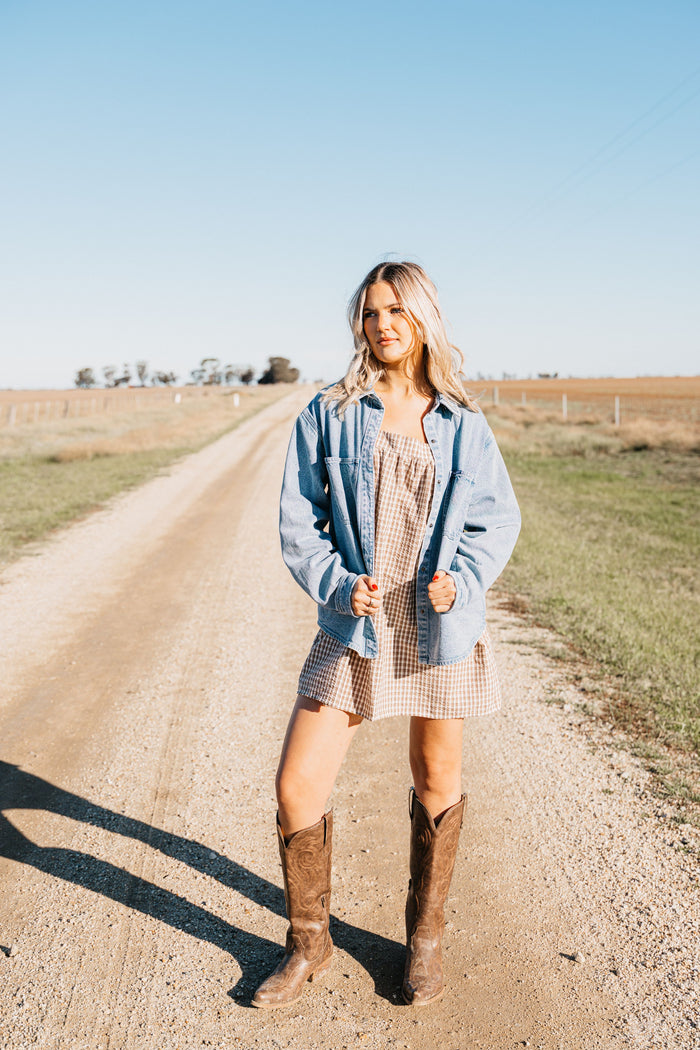 Woman wearing the Lottie Denim Shirt open over a brown gingham dress with cowboy boots on a rural dirt road