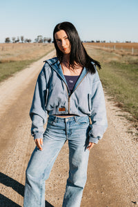Woman wearing a denim jacket and jeans standing on a dirt road with a clear sky.