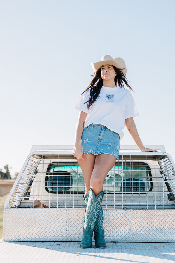 Woman wearing the Montana Tee with a white cowboy hat, light blue embroidered denim skirt and turquoise cowboy boots by a ute