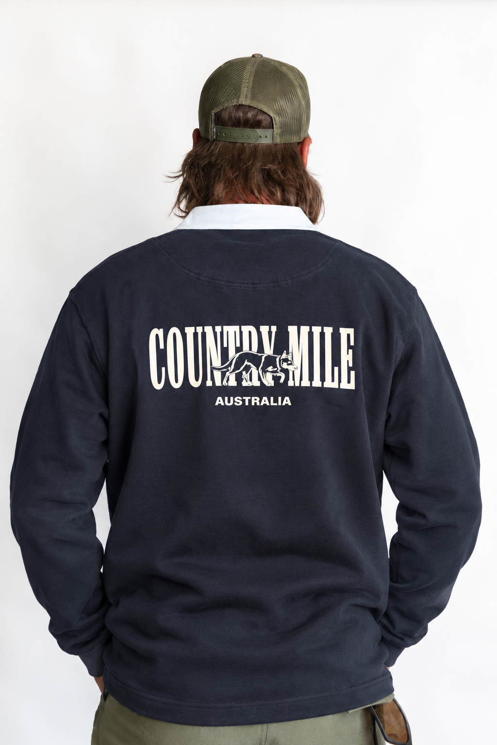 A man viewed from behind wearing a navy blue sweatshirt with a white graphic that reads 'COUNTRY MILE AUSTRALIA' on the back, along with an olive green cap and khaki pants, against a plain white background.