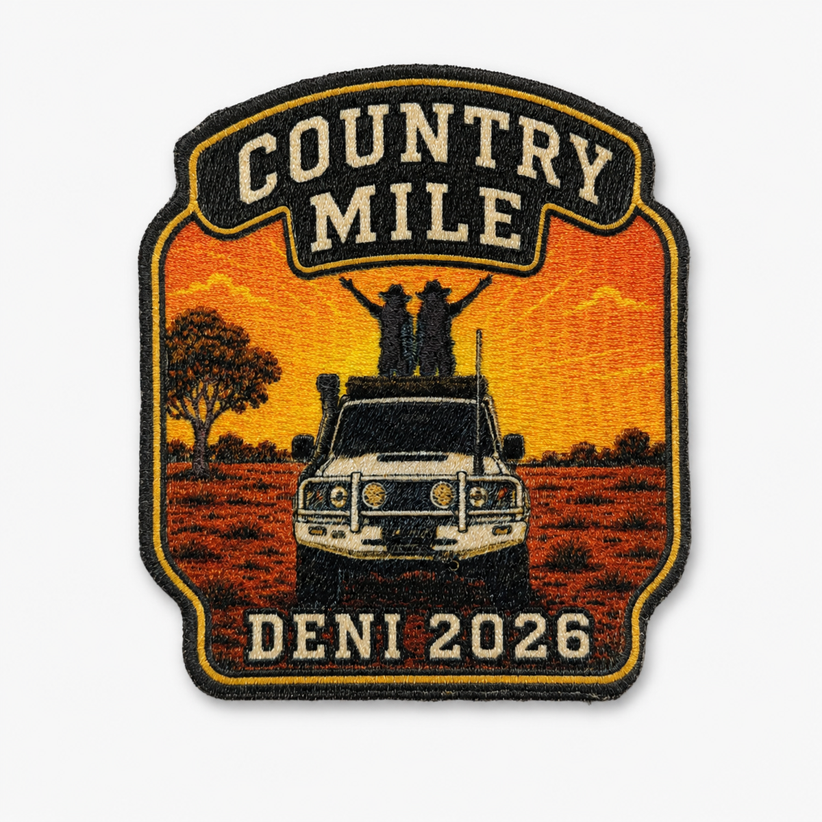 An embroidered patch with a shield-like shape featuring the words 'COUNTRY MILE' at the top and 'DENI 2026' at the bottom. The center shows a white vehicle on an orange field with two people standing on the roof, and a tree on the left, all outlined in black and gold.