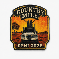 An embroidered patch with a shield-like shape featuring the words 'COUNTRY MILE' at the top and 'DENI 2026' at the bottom. The center shows a white vehicle on an orange field with two people standing on the roof, and a tree on the left, all outlined in black and gold.