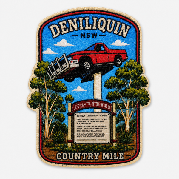 A rectangular embroidered patch with a rounded shield-like border, featuring a red pickup truck jumping over a pole in front of trees and a blue sky. The patch reads “DENILIQUIN -NSW” at the top and “COUNTRY MILE” at the bottom, with a signpost in the center.