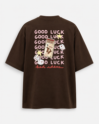 A dark brown oversized short-sleeve t-shirt shown from the back, featuring a graphic print with the word 'GOOD LUCK' repeated in pink, a playing card, dice, and stars.