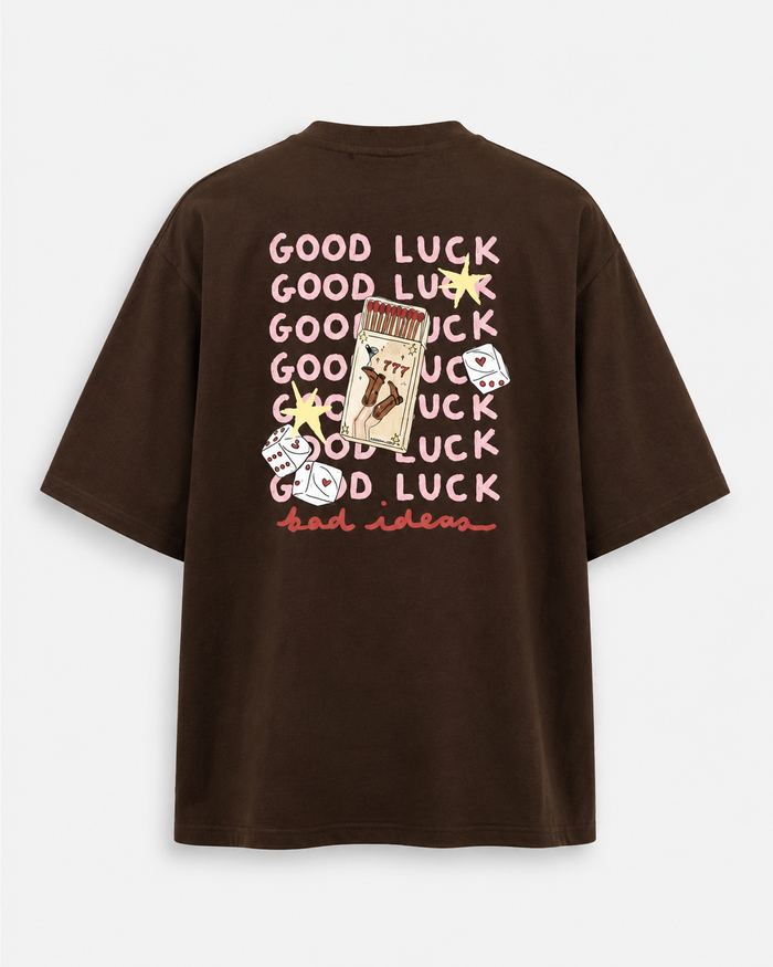 A dark brown oversized short-sleeve t-shirt shown from the back, featuring a graphic print with the word 'GOOD LUCK' repeated in pink, a playing card, dice, and stars.