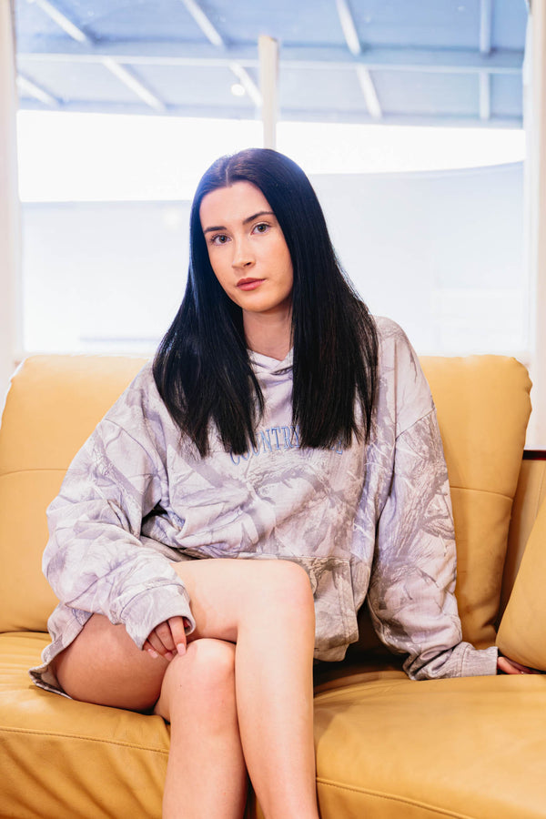 Woman wearing the Grey Camo Hoodie in a light grey bush camo print, seated on a mustard sofa with legs crossed