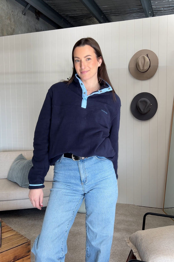Woman wearing the Tilly Fleece in navy with light blue trim, jeans, standing indoors near hats hanging on the wall