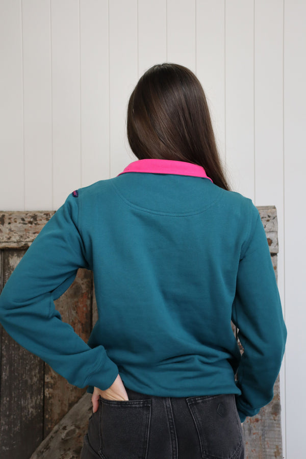 Back view of a woman wearing the Forrest Bonnie Zip in teal with a bright pink collar against a rustic timber wall