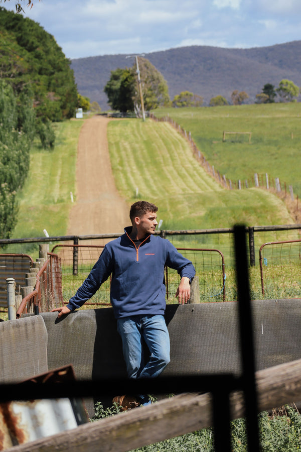 Man wearing the Bradshaw 1/4 Zip in navy with orange zip, leaning on a fence with paddocks and hills in the background