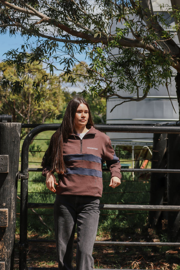 Woman wearing the Bourke Quarter Zip in rich brown with navy chest stripes, standing by a farm gate under trees