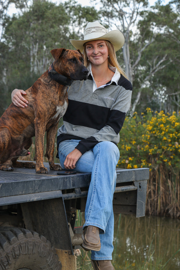 Woman wearing the Flinders Rugby Jumper with jeans and a straw cowboy hat, seated beside a brindle dog outdoors