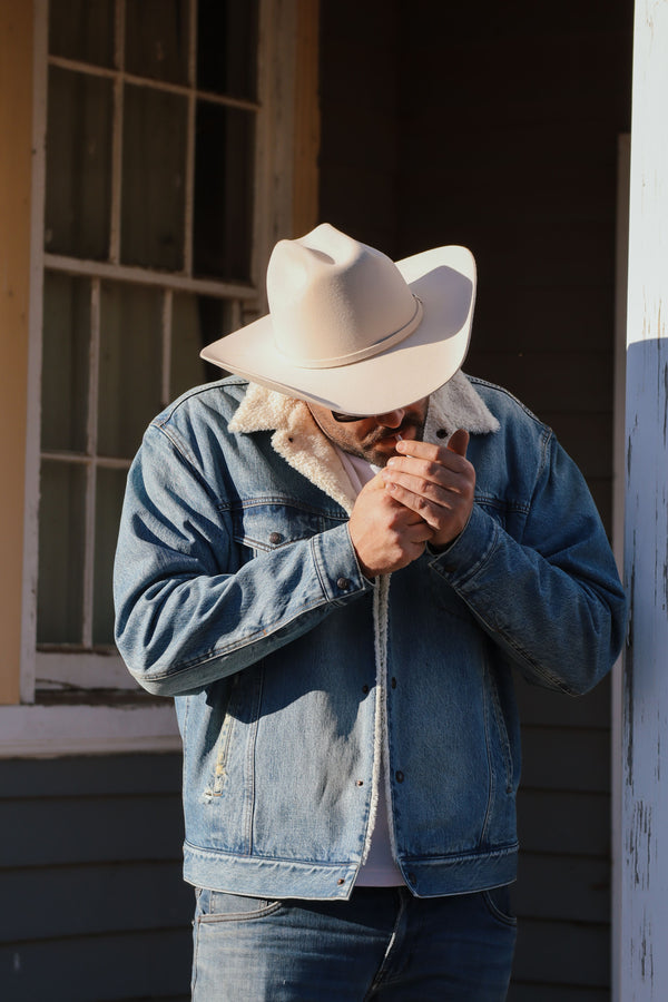 Man wearing the Carter Denim Jacket with a sherpa collar and white cowboy hat, looking down outdoors
