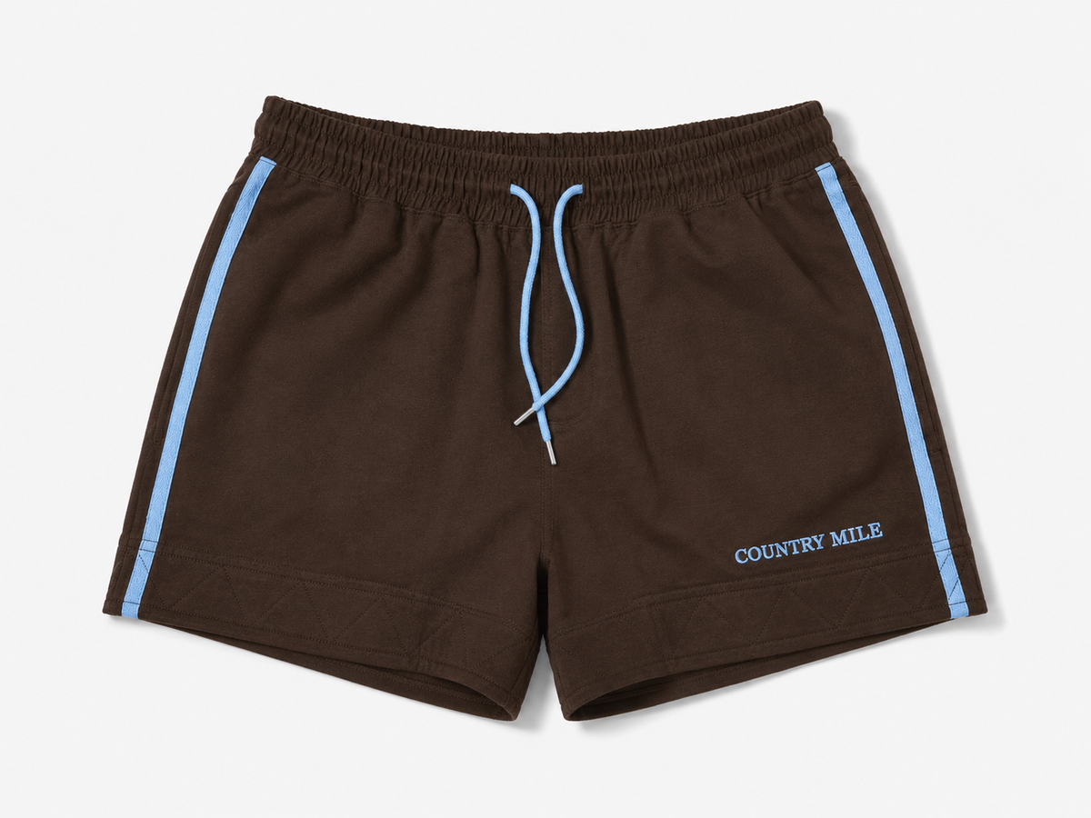 Dark brown work shorts with light blue side stripes, a matching light blue drawstring, and 'COUNTRY MILE' printed in light blue on the lower left leg, shown against a light gray background.