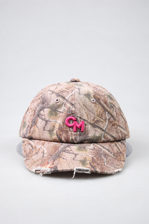 A distressed baseball cap in a brown and beige woodland camouflage pattern, featuring a bright pink embroidered 'CM' logo on the front, photographed against a neutral light gray background.