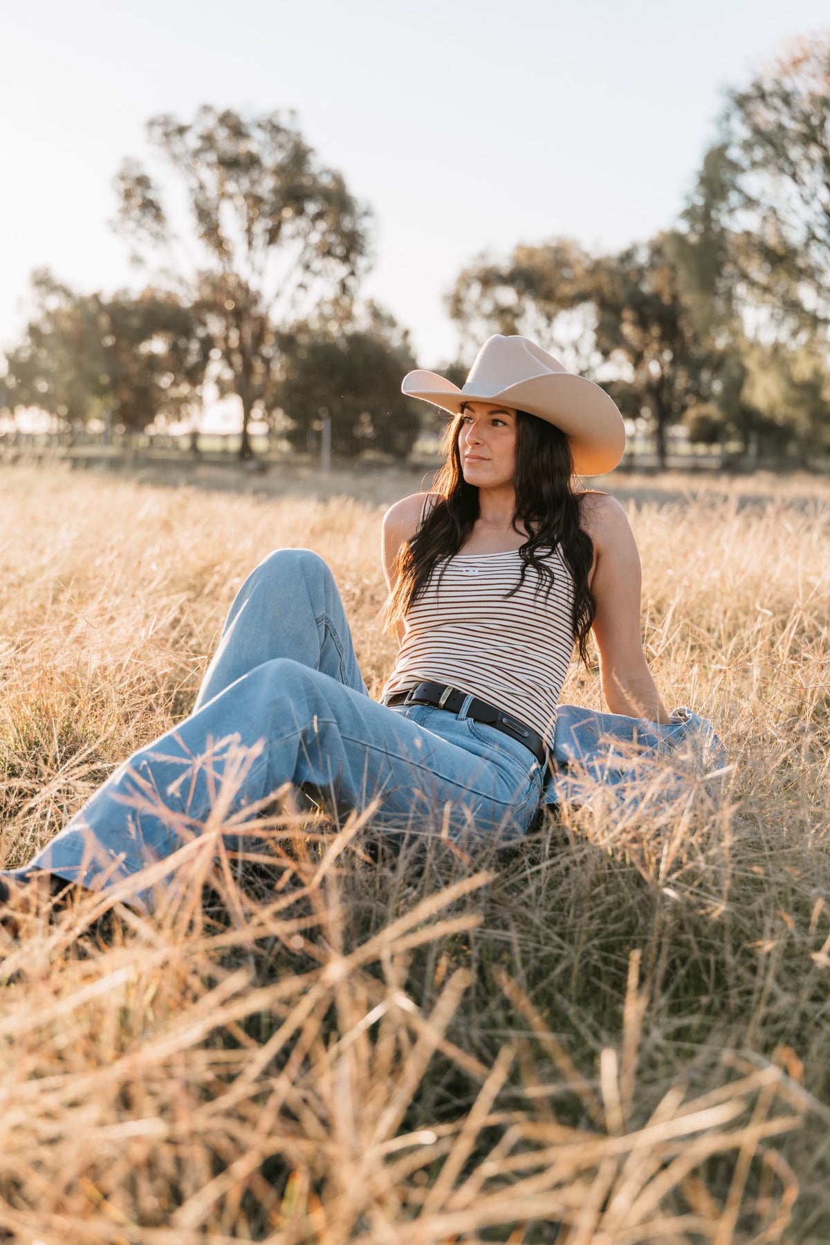 Woman wearing a cowboy hat and striped tank top sitting in a field.