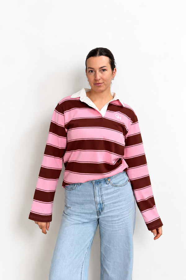 A woman modeling a pink and dark brown striped long-sleeve polo crop top with a white collar, paired with light blue jeans, against a plain white background.