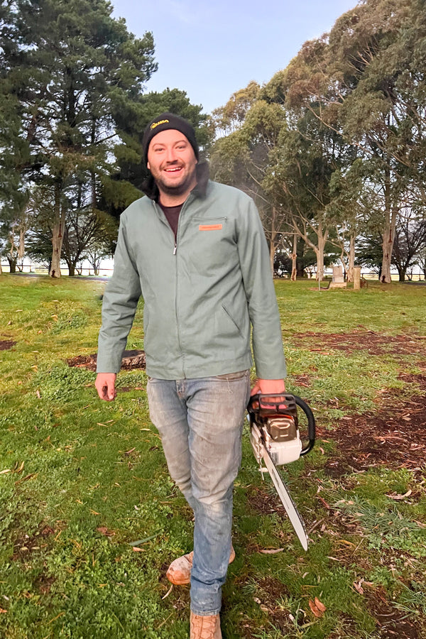 Man wearing the Goulburn Jacket in sage, black beanie, carrying a chainsaw outdoors in a paddock